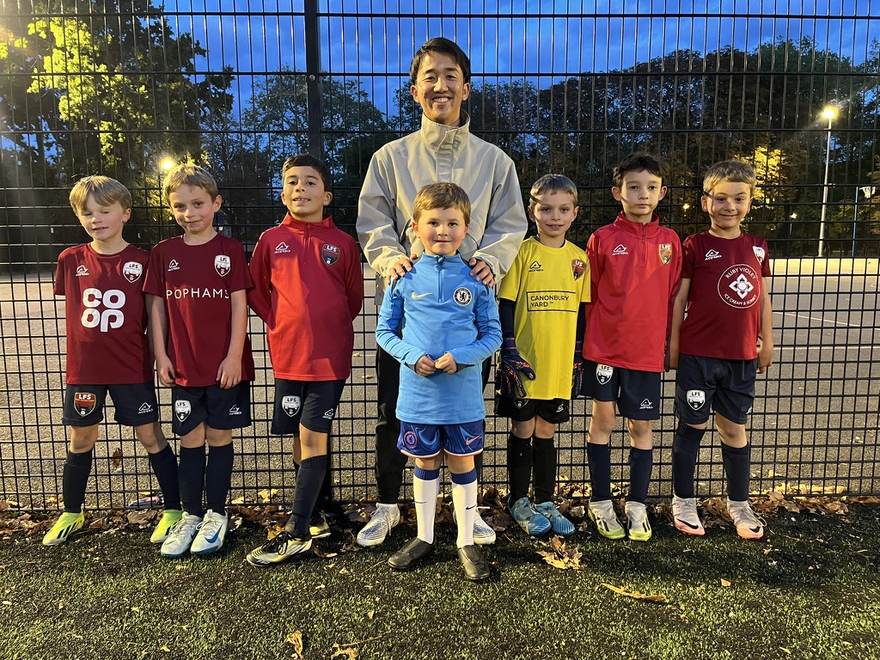 The coach with a group of young players in club kits at a floodlit pitch in London